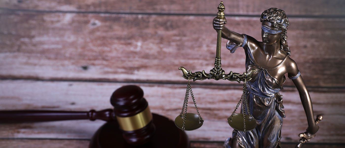 A bronze statue of Lady Justice holding a sword and scale with a gavel in the background.