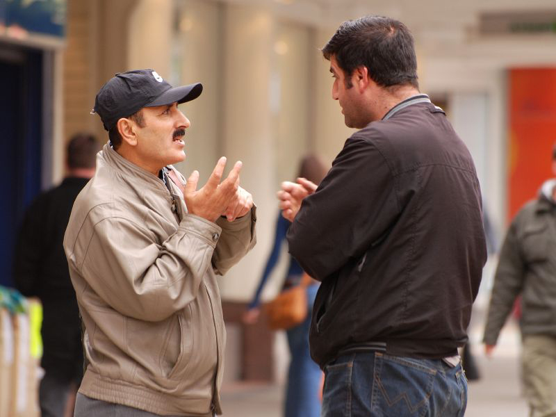 Two people are standing outside. One speaks with his hands open, while the other listens.