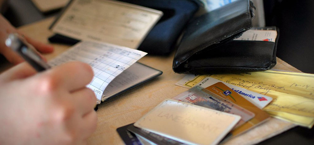 A person's hand is shown writing in a checkbook. Nearby on the table are a wallet, credit cards, and other checks.