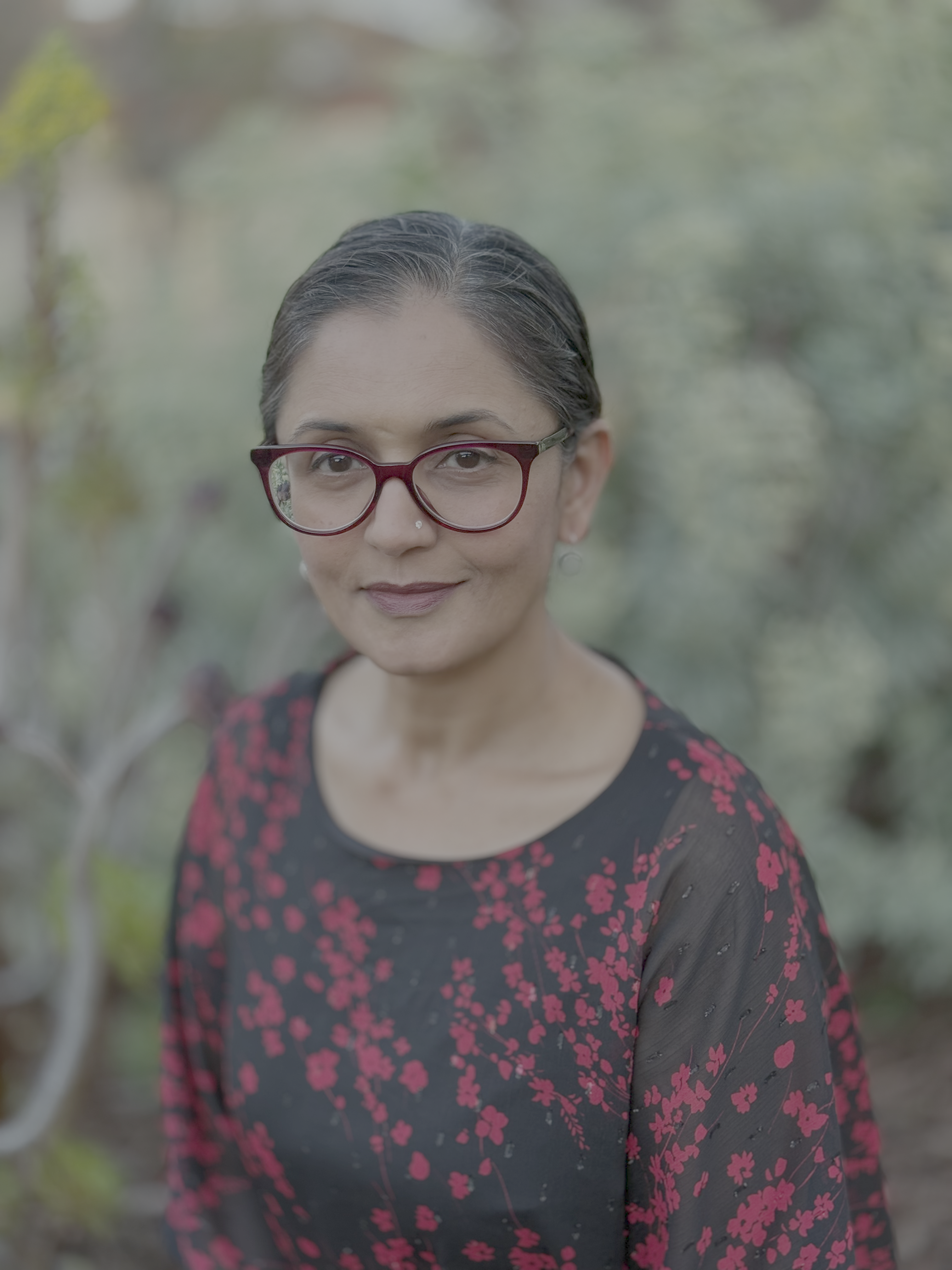 Doli with dark hair pulled back, wearing glasses and a black top with red floral pattern, sitting outdoors.