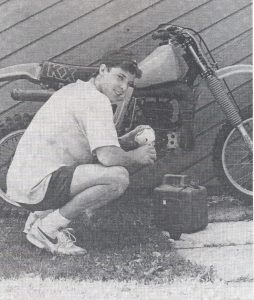 Un joven agachado frente a una motocicleta, llenándola con una mezcla de aceite y gasolina.